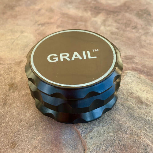Black metal GRAIL™ grinder designed for dried plant material, displayed on a textured brown stone surface