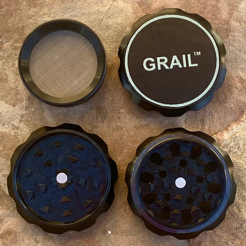 Four-piece GRAIL™ branded metal grinder disassembled to show internal components for processing dried plant material, displayed on a textured brown stone surface
