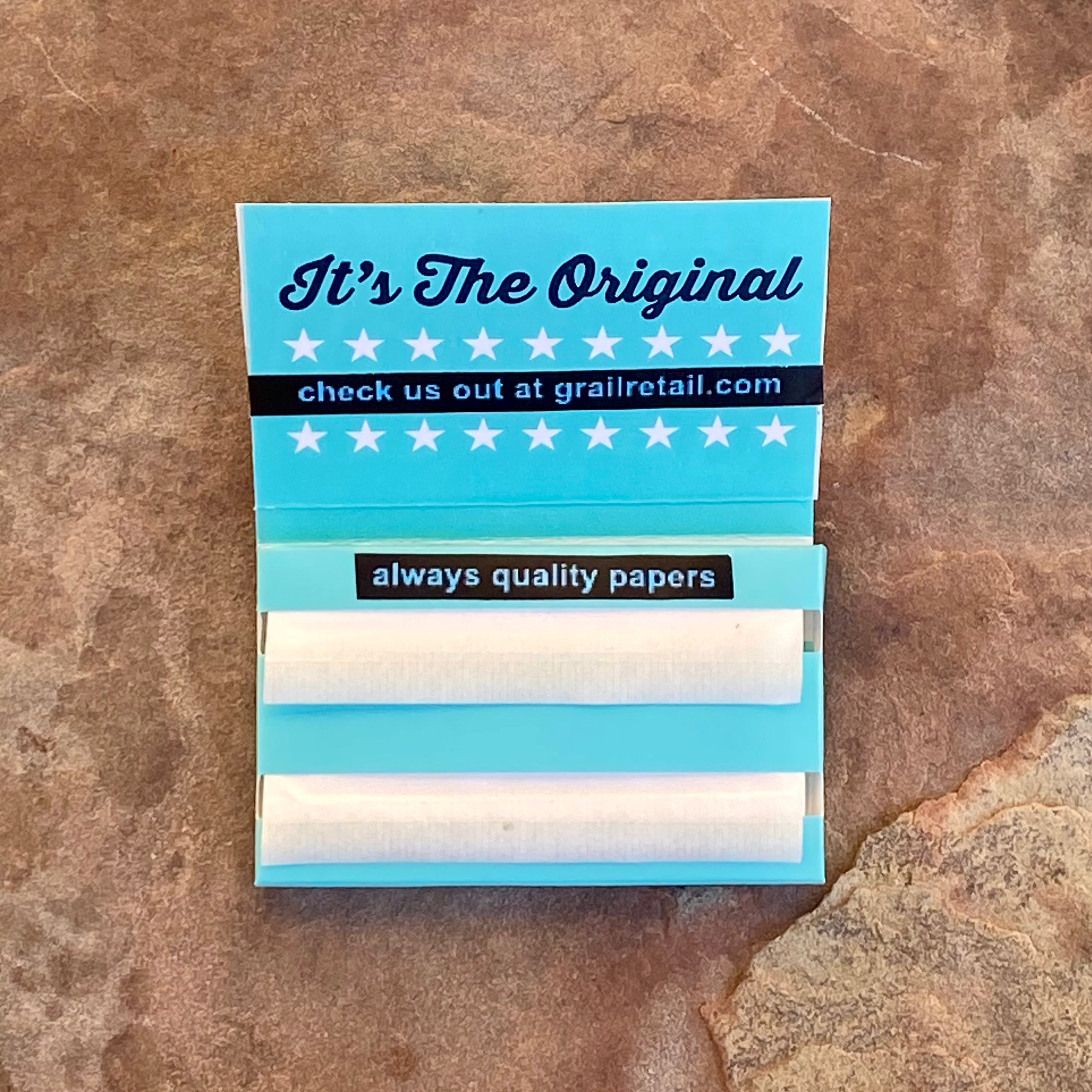 Open pack of Grail™ rolling papers showing individual sheets and inner flap with promotional text, placed on a textured brown stone surface